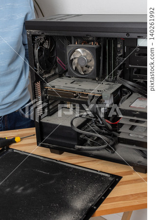 A 53-year-old man removes dust from a computer system unit on a wooden table. He is focused on cleaning the inside of the computer in a well-lit room 140261992
