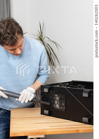 A 53-year-old man uses a vacuum to clean dust from a computer system unit on a wooden table in a home office. The setting is tidy and organized A 53-year-old man uses a vacuum to clean dust from a computer system unit on a wooden table in a home office. The setting is tidy and organized 140262003