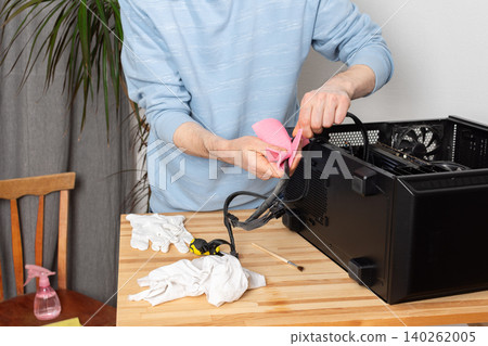 A 53-year-old man uses a cloth to clean dust from a computer system unit. He works at a wooden table in a room with plants nearby. Tools are set out for the task A 53-year-old man uses a cloth to clean dust from a computer system unit. He works at a wooden table in a room with plants nearby. Tools are set out for the task 140262005