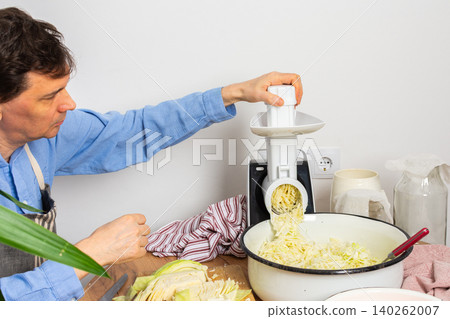 A 53-year-old man prepares cabbage and carrots for fermentation in his kitchen. He uses a machine to shred the vegetables while surrounded by kitchen tools and ingredients A 53-year-old man prepares cabbage and carrots for fermentation in his kitchen. He uses a machine to shred the vegetables while surrounded by kitchen tools and ingredients 140262007