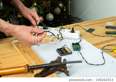 A man fixes a Christmas tree garland and lights on a table at home. He is checking the connection and making adjustments to ensure everything works for the holiday season 140262013