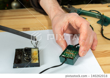 A man works on fixing a broken Christmas tree garland by soldering wires together. He uses tools and parts on a wooden table, focusing on the repair process during the holiday season A man works on fixing a broken Christmas tree garland by soldering wires together. He uses tools and parts on a wooden table, focusing on the repair process during the holiday season 140262018