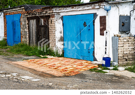 An Abandoned Garage Scene Featuring Blue Doors, Rustic Walls, and a Vibrant Carpet on a Gravel Path Within a Green, Overgrown Environment An Abandoned Garage Scene Featuring Blue Doors, Rustic Walls, and a Vibrant Carpet on a Gravel Path Within a Green, Overgrown Environment 140262032