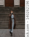 Woman walks on steps wearing a long coat and dress in a city location during the day 140262196