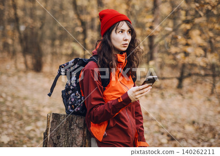 Woman looks away while holding smartphone and leaning against tree stump in forest. solo female travel safety, self-reliant outdoor exploration, wilderness preparedness. Woman looks away while holding smartphone and leaning against tree stump in forest. solo female travel safety, self-reliant outdoor exploration, wilderness preparedness. 140262211
