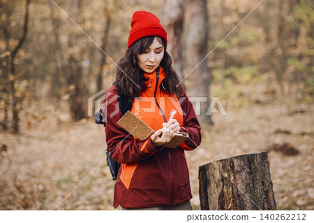 Woman stands in autumn forest and writes in notebook while leaning on tree stump. Woman forest inspector, female industrial professional, environmental auditor at work. Woman stands in autumn forest and writes in notebook while leaning on tree stump. Woman forest inspector, female industrial professional, environmental auditor at work. 140262212