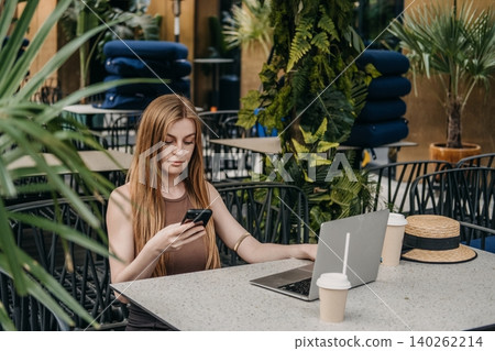 Young woman sits at outdoor cafe terrace looking at smartphone with open laptop on table. Social media manager, influencer content creation, mobile business application. Young woman sits at outdoor cafe terrace looking at smartphone with open laptop on table. Social media manager, influencer content creation, mobile business application. 140262214
