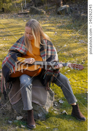 Beautiful blonde girl playing acoustic guitar and singing relaxing enjoying outdoors on sunny autumn day. Hippie woman in boho style learning and practicing playing musical instrument. Fall leaves 140262245