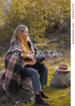 Blonde woman playing string guitar outdoors in autumn forest. Concept of sound therapy, mental health and wellness rituals. Calmness tranquility audio-sensory practices. Aura farming energy 140262254