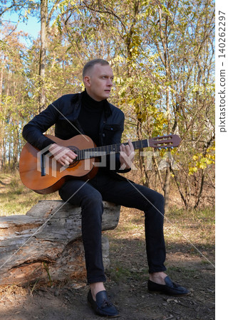 Portrait of smiling caucasian artist plays wooden acoustic guitar in autumnal park. Young songwriter plays string musical instrument hobby outside in nature fall time. Audio music healing Portrait of smiling caucasian artist plays wooden acoustic guitar in autumnal park. Young songwriter plays string musical instrument hobby outside in nature fall time. Audio music healing 140262297