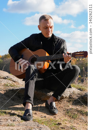 Portrait of smiling caucasian artist plays wooden acoustic guitar in autumnal park. Young songwriter plays string musical instrument hobby outside in nature fall time. Audio music healing Portrait of smiling caucasian artist plays wooden acoustic guitar in autumnal park. Young songwriter plays string musical instrument hobby outside in nature fall time. Audio music healing 140262311