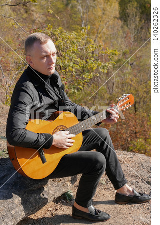 Portrait of smiling caucasian artist plays wooden acoustic guitar in autumnal park. Young songwriter plays string musical instrument hobby outside in nature fall time. Audio music healing Portrait of smiling caucasian artist plays wooden acoustic guitar in autumnal park. Young songwriter plays string musical instrument hobby outside in nature fall time. Audio music healing 140262326