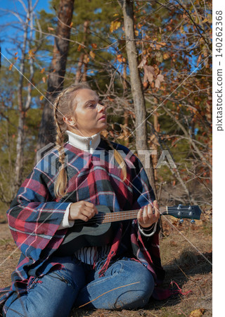 Beautiful blonde girl playing guitar ukulele and singing relaxing enjoying outdoors on sunny autumn day. Hippie woman in boho style training playing musical instrument. Fall leaves Beautiful blonde girl playing guitar ukulele and singing relaxing enjoying outdoors on sunny autumn day. Hippie woman in boho style training playing musical instrument. Fall leaves 140262368