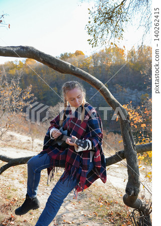 Beautiful blonde girl playing guitar ukulele and singing relaxing enjoying outdoors on sunny autumn day. Hippie woman in boho style training playing musical instrument. Fall leaves 140262415