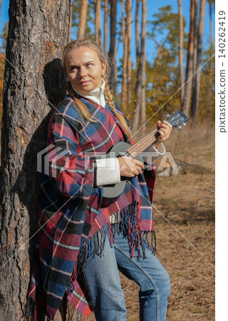 Blonde woman playing guitar ukulele outdoors in autumn forest. Concept of sound therapy, mental health and wellness rituals. Calmness tranquility audio-sensory practices. Aura farming Blonde woman playing guitar ukulele outdoors in autumn forest. Concept of sound therapy, mental health and wellness rituals. Calmness tranquility audio-sensory practices. Aura farming 140262419