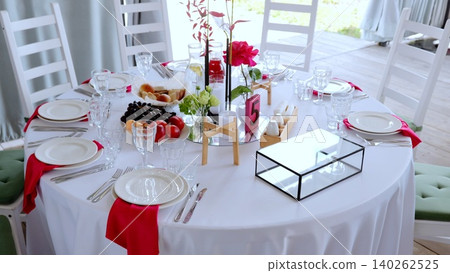 Elegant round dining setup with crisp white tablecloth, pristine dinnerware, sparkling glassware, and vibrant red rose centerpiece enhancing celebratory table design 140262525