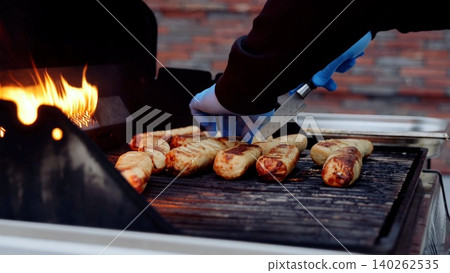 Street food chef wearing blue gloves grilling sausages over flaming barbecue, carefully monitoring cooking process for optimal flavor Street food chef wearing blue gloves grilling sausages over flaming barbecue, carefully monitoring cooking process for optimal flavor 140262535
