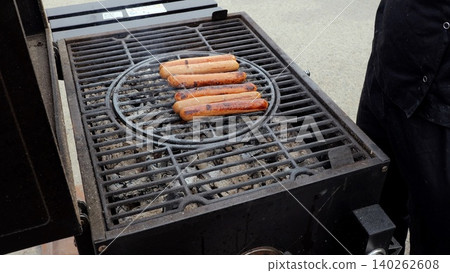 Festivals feature open flame sausage grilling, Sizzling sausages and smoky scents enchant festival attendees, Chef monitors sausages roasting on open grill amid smoky aroma at outdoor festival Festivals feature open flame sausage grilling, Sizzling sausages and smoky scents enchant festival attendees, Chef monitors sausages roasting on open grill amid smoky aroma at outdoor festival 140262608