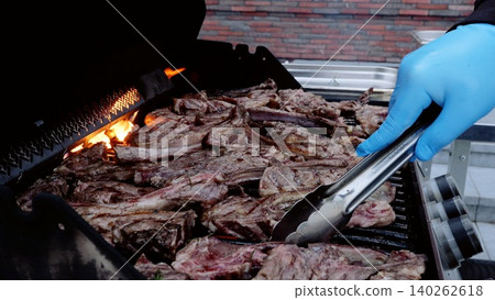 Chef in blue gloves grilling lamb ribs over flaming barbecue during outdoor catering preparation Chef in blue gloves grilling lamb ribs over flaming barbecue during outdoor catering preparation 140262618