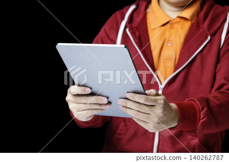 Person in red hoodie holding digital tablet. Dark background highlights device and gesture. User appears engaged, attentive to screen. Ideal for education, business. High quality photo 140262787