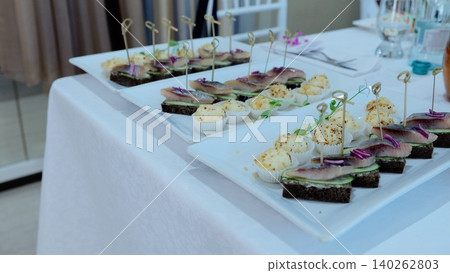 Herring and cheese mini sandwiches arranged neatly on pristine white tablecloth, presenting elegant appetizer selection for sophisticated catering event Herring and cheese mini sandwiches arranged neatly on pristine white tablecloth, presenting elegant appetizer selection for sophisticated catering event 140262803