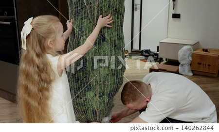 Father and daughter carefully unwrapping festive christmas tree from protective packaging, sharing warm holiday moment together in comfortable living space Father and daughter carefully unwrapping festive christmas tree from protective packaging, sharing warm holiday moment together in comfortable living space 140262844