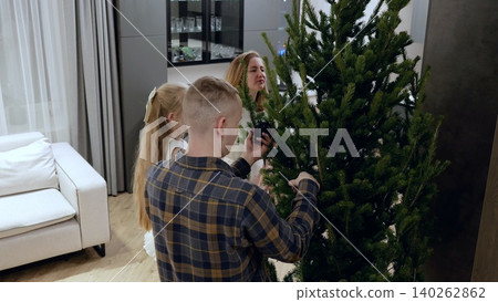 Family gathering around sparkling christmas tree, hanging ornaments, sharing laughter in contemporary living space filled with warmth and seasonal cheer Family gathering around sparkling christmas tree, hanging ornaments, sharing laughter in contemporary living space filled with warmth and seasonal cheer 140262862