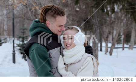 Affectionate couple hugging tenderly, wearing winter coats and fluffy earmuffs, standing amid snow covered trees in picturesque park landscape Affectionate couple hugging tenderly, wearing winter coats and fluffy earmuffs, standing amid snow covered trees in picturesque park landscape 140262908