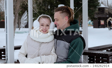 Winter park gazebo framing smiling young couple, wearing warm clothing, embracing snowy landscape with bare trees and soft white scenery surrounding happy partners Winter park gazebo framing smiling young couple, wearing warm clothing, embracing snowy landscape with bare trees and soft white scenery surrounding happy partners 140262914