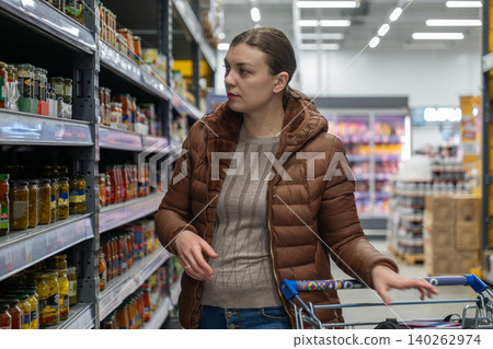 Woman in supermarket aisle reaching for top shelf item, determined lastminute shopping with focused gaze, brown padded coat, trolley beside, neatly stacked goods and bright overhead lighting, brisk Woman in supermarket aisle reaching for top shelf item, determined lastminute shopping with focused gaze, brown padded coat, trolley beside, neatly stacked goods and bright overhead lighting, brisk 140262974