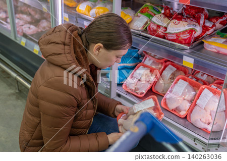 Woman reading frozen chicken label closely, checking ingredients and expiry while holding cart handle at refrigerated display, focused expression, prepping weeknight dinner and comparing value Woman reading frozen chicken label closely, checking ingredients and expiry while holding cart handle at refrigerated display, focused expression, prepping weeknight dinner and comparing value 140263036
