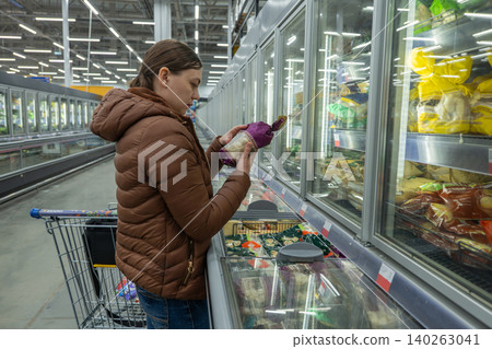 Woman in supermarket comparing prices and labels, scanning frozen items for best value while leaning on cart practical budgeting mood, focused posture, retail signage visible, decisionmaking moment 140263041