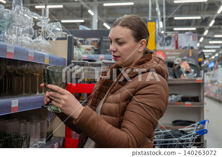 Caucasian Woman Inspecting Glass Vase In Store Aisle, Wearing Brown Puffer Jacket, Holding Green Glass, Shopping Cart Visible, Fluorescent Lighting, Focused Expression, Comparing Textures And Price. Caucasian Woman Inspecting Glass Vase In Store Aisle, Wearing Brown Puffer Jacket, Holding Green Glass, Shopping Cart Visible, Fluorescent Lighting, Focused Expression, Comparing Textures And Price. 140263072