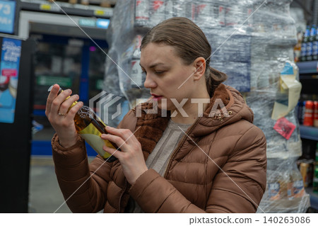 White woman comparing discounted beer price near checkout, focused expression, budget conscious purchase, stacked pallets and promotional tags visible, casual jacket and artificial lighting White woman comparing discounted beer price near checkout, focused expression, budget conscious purchase, stacked pallets and promotional tags visible, casual jacket and artificial lighting 140263086