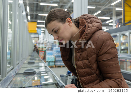 Woman in supermarket selecting frozen meal for healthy dinner, reading ingredients and nutrition facts, intent shopper in brown jacket, chilled aisle with packaged options, mindful food decision Woman in supermarket selecting frozen meal for healthy dinner, reading ingredients and nutrition facts, intent shopper in brown jacket, chilled aisle with packaged options, mindful food decision 140263135