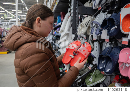 White Woman Holding Red Sandal Pair Against Wall Display, Casual Brown Coat, Comparing Size And Strap, Bustling Superstore Aisle, Shopping Cart Nearby, Colorful Shoe Rows, Focused Selection For Summer White Woman Holding Red Sandal Pair Against Wall Display, Casual Brown Coat, Comparing Size And Strap, Bustling Superstore Aisle, Shopping Cart Nearby, Colorful Shoe Rows, Focused Selection For Summer 140263148