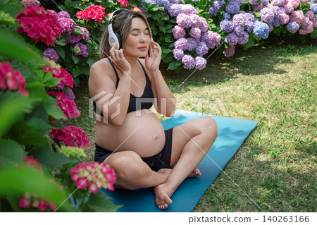 Pregnant asian woman sitting cross legged on a yoga mat in a garden with hydrangeas, listening to music through headphones, her eyes closed in peaceful meditation.Music therapy Pregnant asian woman sitting cross legged on a yoga mat in a garden with hydrangeas, listening to music through headphones, her eyes closed in peaceful meditation.Music therapy 140263166