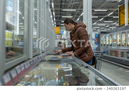 Woman in supermarket selecting frozen meals for meal prep, scanning packaging for cooking instructions and nutrition while holding cart practical evening planning vibe, refrigerated display Woman in supermarket selecting frozen meals for meal prep, scanning packaging for cooking instructions and nutrition while holding cart practical evening planning vibe, refrigerated display 140263197