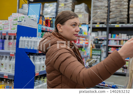 Woman Selecting Shower Sponge In Store, Focused Expression Scanning Texture And Label, Brown Puffer Jacket, Packed Shelves Of Toiletries, Fluorescent Lighting, Everyday Errand Mood. Woman Selecting Shower Sponge In Store, Focused Expression Scanning Texture And Label, Brown Puffer Jacket, Packed Shelves Of Toiletries, Fluorescent Lighting, Everyday Errand Mood. 140263232