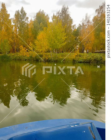 Autumn landscape with colorful trees reflecting in calm water, viewed from a blue boat on a serene lake surrounded by vibrant foliage and cloudy sky Autumn landscape with colorful trees reflecting in calm water, viewed from a blue boat on a serene lake surrounded by vibrant foliage and cloudy sky 140263254