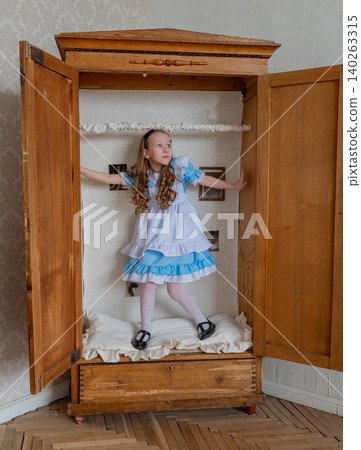 Young Girl Inside Wooden Wardrobe Playing Hideandseek, Wearing Blue Ruffled Dress, Joyful Expression, Soft Natural Light, Quilted Lining, Antique Furniture, Parquet Floor, Floral Wallpaper. Young Girl Inside Wooden Wardrobe Playing Hideandseek, Wearing Blue Ruffled Dress, Joyful Expression, Soft Natural Light, Quilted Lining, Antique Furniture, Parquet Floor, Floral Wallpaper. 140263315