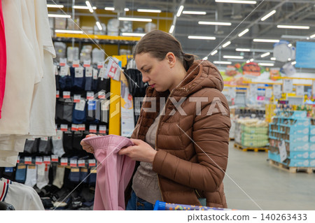 White Woman Evaluating Pink Top On Hanger, Brown Coat, Scanning Size And Label Under Store Lights, Shopping Cart Nearby, Thoughtful Expression, Seasonal Wardrobe Update, Instore Discovery 140263433