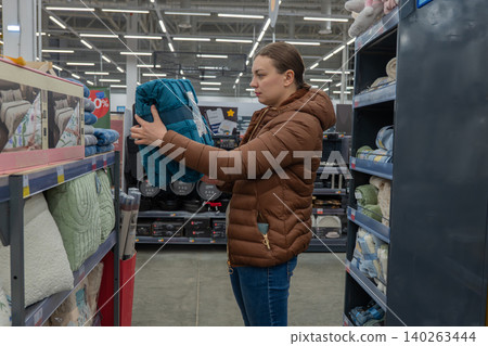Customer Selecting Fabric In Store. Woman Evaluates Teal Throw While Browsing Crowded Shop Aisles. Female Shopper Carefully Examines Teal Blanket Among Many Other Household Items In Store Aisle 140263444