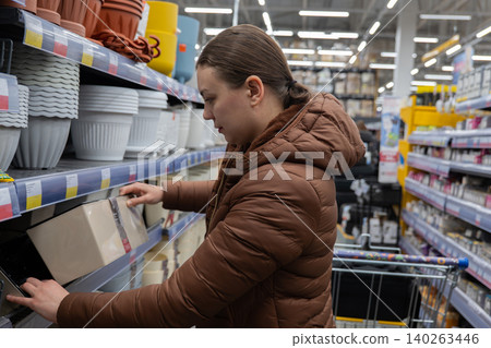 White Woman Inspecting Kitchen Tray Shelves, Brown Puffer Coat, Shopping Cart, Fluorescent Superstore Lighting, Rows Of Planters And Storage Containers, Checking Texture And Price Label, Focused White Woman Inspecting Kitchen Tray Shelves, Brown Puffer Coat, Shopping Cart, Fluorescent Superstore Lighting, Rows Of Planters And Storage Containers, Checking Texture And Price Label, Focused 140263446
