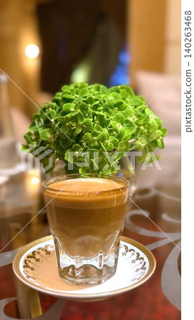 Glass latte stands beside fresh green blossoms in warm cafe ambiance, showcasing creamy coffee texture, elegant table styling, cozy hospitality, travel destination charm, and latte lifestyle. Glass latte stands beside fresh green blossoms in warm cafe ambiance, showcasing creamy coffee texture, elegant table styling, cozy hospitality, travel destination charm, and latte lifestyle. 140263468