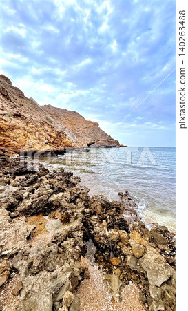 Rocky coastal cliffs curve beside gentle water under bright sky, highlighting rugged shoreline textures, remote natural beauty, peaceful seascape, and travel destination value along coastal scenery. Rocky coastal cliffs curve beside gentle water under bright sky, highlighting rugged shoreline textures, remote natural beauty, peaceful seascape, and travel destination value along coastal scenery. 140263488