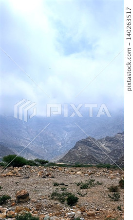 Mountain landscape spreads beneath pale cloudy sky with rocky foreground, mountain ridges, desert shrubs, soft haze, and travel destination atmosphere for rugged nature scenery. Mountain landscape spreads beneath pale cloudy sky with rocky foreground, mountain ridges, desert shrubs, soft haze, and travel destination atmosphere for rugged nature scenery. 140263517