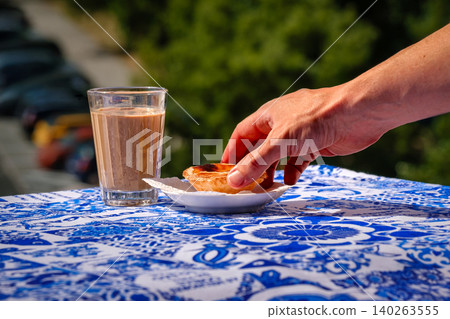 Portuguese custard tart with a hand reaching near coffee on a blue table beside a white plate with blurred background. Traditional pastry, culinary travel, authentic taste, destination dessert Portuguese custard tart with a hand reaching near coffee on a blue table beside a white plate with blurred background. Traditional pastry, culinary travel, authentic taste, destination dessert 140263555