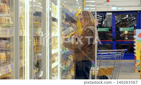 Woman in brown leather jacket choosing frozen food from display case filled with cakes and pastries in well lit supermarket 140263567