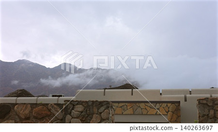 Cloudy Mountain View. Foggy Peaks Behind Quiet Roof. Silent Skyline With Distant Mountain Silhouettes. Serene Horizon Framed By Modern Building And Misty Clouds 140263697